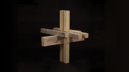 Kreuzförmige Holzverbindung aus hellen Balken mit einem dunkleren Altholzbalken, vor schwarzem Hintergrund.