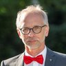 "Porträtfotografie eines älteren Mannes mit grauem Haar und Brille vor grünem unscharfem Hintergrund. Er trägt einen grauen Anzug, weißes Hemd, rote Fliege und rotes Einstecktuch. Sein Blick richtet sich direkt zur Kamera, sein Gesichtsausdruck wirkt ruhig und professionell."