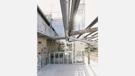 Innenhof mit Glasaufzug und Stegen; darüber verlaufende große Metallrohre, Sonnenstrahlen blenden durch die Konstruktion.