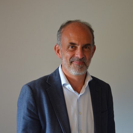 Porträtfoto eines älteren Mannes mit grauem Haar und Bart, der einen dunkelblauen Blazer über einem hellblauen Hemd trägt. Er lächelt leicht in die Kamera vor neutralem grauem Hintergrund."