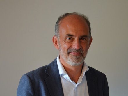Porträtfoto eines älteren Mannes mit grauem Haar und Bart, der einen dunkelblauen Blazer über einem hellblauen Hemd trägt. Er lächelt leicht in die Kamera vor neutralem grauem Hintergrund."