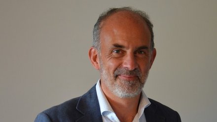 Porträtfoto eines älteren Mannes mit grauem Haar und Bart, der einen dunkelblauen Blazer über einem hellblauen Hemd trägt. Er lächelt leicht in die Kamera vor neutralem grauem Hintergrund."