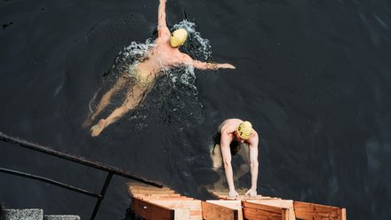 Zwei Personen mit Badekappen im dunklen Wasser; eine schwimmt Kraul, die andere steigt über eine Holztreppe aus dem Wasser.