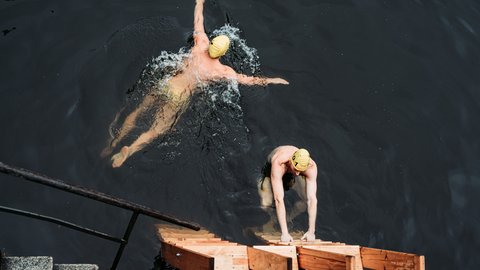 Zwei Personen mit Badekappen im dunklen Wasser; eine schwimmt Kraul, die andere steigt über eine Holztreppe aus dem Wasser.