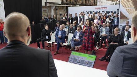 Publikum bei einer Podiumsdiskussion; eine Frau spricht mit Mikrofon, andere hören zu.