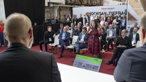 Publikum bei einer Podiumsdiskussion; eine Frau spricht mit Mikrofon, andere hören zu.