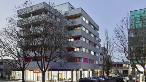 "Sechsgeschossiges modernes Wohngebäude mit farbigen Balkonen in Rot und Orange, Ladengeschäft im Erdgeschoss, geparkter Autos davor, kahl Bäume, Dämmerung."