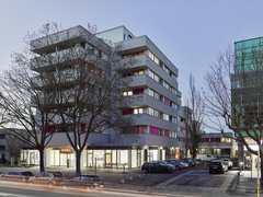 "Sechsgeschossiges modernes Wohngebäude mit farbigen Balkonen in Rot und Orange, Ladengeschäft im Erdgeschoss, geparkter Autos davor, kahl Bäume, Dämmerung."