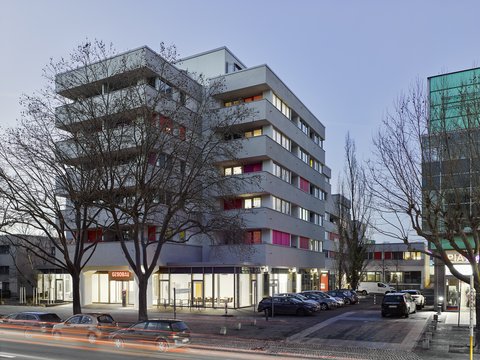 "Sechsgeschossiges modernes Wohngebäude mit farbigen Balkonen in Rot und Orange, Ladengeschäft im Erdgeschoss, geparkter Autos davor, kahl Bäume, Dämmerung."