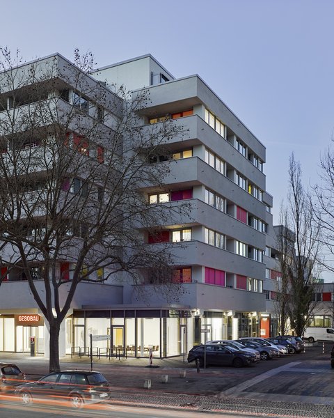 "Sechsgeschossiges modernes Wohngebäude mit farbigen Balkonen in Rot und Orange, Ladengeschäft im Erdgeschoss, geparkter Autos davor, kahl Bäume, Dämmerung."