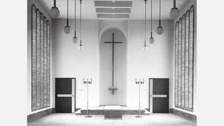 Schwarzweiß-Fotografie eines minimalistischen Kirchenraums mit weißen Wänden. In der Mitte hängt ein großes Kreuz an der Wand über einem Altar mit Stufen. Gegenüber sind zwei schwarze Türen, rechts und links hängen strukturierte Vorhänge. Von der Decke hängen mehrere Pendelleuchten mit Kugeln. Der Raum verkörpert moderne protestantische Kirchenarchitektur.