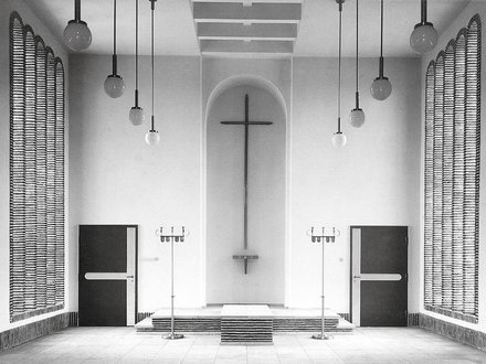 Schwarzweiß-Fotografie eines minimalistischen Kirchenraums mit weißen Wänden. In der Mitte hängt ein großes Kreuz an der Wand über einem Altar mit Stufen. Gegenüber sind zwei schwarze Türen, rechts und links hängen strukturierte Vorhänge. Von der Decke hängen mehrere Pendelleuchten mit Kugeln. Der Raum verkörpert moderne protestantische Kirchenarchitektur.