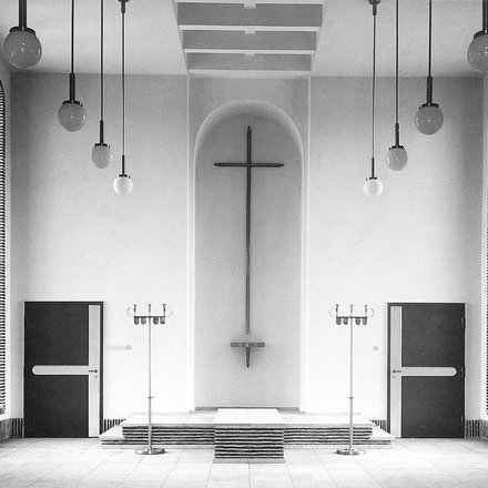 Schwarzweiß-Fotografie eines minimalistischen Kirchenraums mit weißen Wänden. In der Mitte hängt ein großes Kreuz an der Wand über einem Altar mit Stufen. Gegenüber sind zwei schwarze Türen, rechts und links hängen strukturierte Vorhänge. Von der Decke hängen mehrere Pendelleuchten mit Kugeln. Der Raum verkörpert moderne protestantische Kirchenarchitektur.