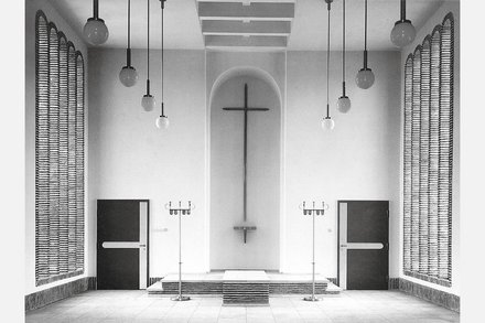 Schwarzweiß-Fotografie eines minimalistischen Kirchenraums mit weißen Wänden. In der Mitte hängt ein großes Kreuz an der Wand über einem Altar mit Stufen. Gegenüber sind zwei schwarze Türen, rechts und links hängen strukturierte Vorhänge. Von der Decke hängen mehrere Pendelleuchten mit Kugeln. Der Raum verkörpert moderne protestantische Kirchenarchitektur.