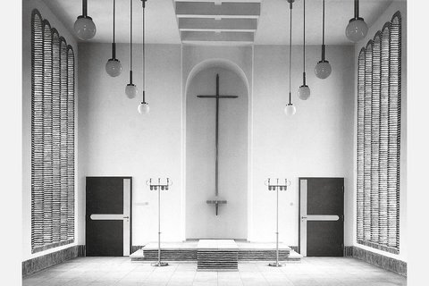 Schwarzweiß-Fotografie eines minimalistischen Kirchenraums mit weißen Wänden. In der Mitte hängt ein großes Kreuz an der Wand über einem Altar mit Stufen. Gegenüber sind zwei schwarze Türen, rechts und links hängen strukturierte Vorhänge. Von der Decke hängen mehrere Pendelleuchten mit Kugeln. Der Raum verkörpert moderne protestantische Kirchenarchitektur.