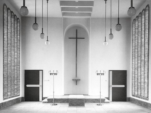 Schwarzweiß-Fotografie eines minimalistischen Kirchenraums mit weißen Wänden. In der Mitte hängt ein großes Kreuz an der Wand über einem Altar mit Stufen. Gegenüber sind zwei schwarze Türen, rechts und links hängen strukturierte Vorhänge. Von der Decke hängen mehrere Pendelleuchten mit Kugeln. Der Raum verkörpert moderne protestantische Kirchenarchitektur.