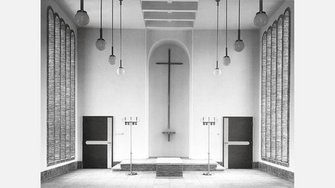 Schwarzweiß-Fotografie eines minimalistischen Kirchenraums mit weißen Wänden. In der Mitte hängt ein großes Kreuz an der Wand über einem Altar mit Stufen. Gegenüber sind zwei schwarze Türen, rechts und links hängen strukturierte Vorhänge. Von der Decke hängen mehrere Pendelleuchten mit Kugeln. Der Raum verkörpert moderne protestantische Kirchenarchitektur.