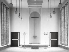 Schwarzweiß-Fotografie eines minimalistischen Kirchenraums mit weißen Wänden. In der Mitte hängt ein großes Kreuz an der Wand über einem Altar mit Stufen. Gegenüber sind zwei schwarze Türen, rechts und links hängen strukturierte Vorhänge. Von der Decke hängen mehrere Pendelleuchten mit Kugeln. Der Raum verkörpert moderne protestantische Kirchenarchitektur.