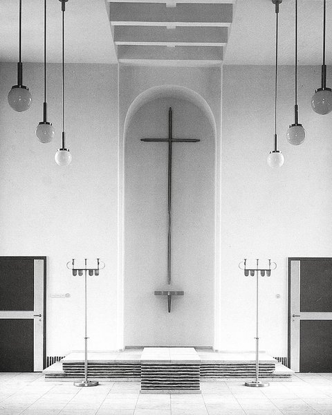 Schwarzweiß-Fotografie eines minimalistischen Kirchenraums mit weißen Wänden. In der Mitte hängt ein großes Kreuz an der Wand über einem Altar mit Stufen. Gegenüber sind zwei schwarze Türen, rechts und links hängen strukturierte Vorhänge. Von der Decke hängen mehrere Pendelleuchten mit Kugeln. Der Raum verkörpert moderne protestantische Kirchenarchitektur.
