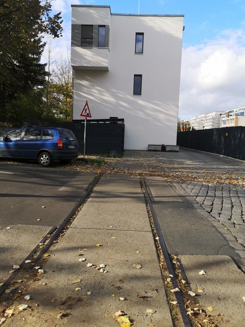Straße mit Straßenbahngleisen im Vordergrund und einem modernen, weißen Wohngebäude; am Straßenrand steht ein Baustellenschild.