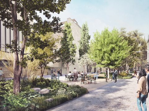 Gestalteter öffentlicher Platz mit alten Bäumen, Sitzgruppen aus Stühlen und Tischen. Menschen spazieren und sitzen im Schatten. Terrakottafarbene und beige Gebäude im Hintergrund. Gepflasterte Fläche, grüne Pflanzen und Büsche.