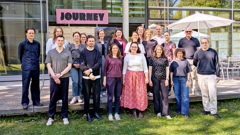 "Gruppenfoto von etwa 25 Personen verschiedenen Alters und Geschlechts, die nebeneinander auf einer Wiese vor einem modernen Gebäude mit großen Glasfenstern stehen. Über dem Eingang ein rosafarbenes Schild mit 'JOURNEY'-Aufschrift. Ein weißer Sonnenschirm ist rechts sichtbar. Klare Herbsttag mit bewölktem Himmel."