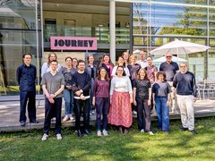 "Gruppenfoto von etwa 25 Personen verschiedenen Alters und Geschlechts, die nebeneinander auf einer Wiese vor einem modernen Gebäude mit großen Glasfenstern stehen. Über dem Eingang ein rosafarbenes Schild mit 'JOURNEY'-Aufschrift. Ein weißer Sonnenschirm ist rechts sichtbar. Klare Herbsttag mit bewölktem Himmel."