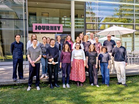 "Gruppenfoto von etwa 25 Personen verschiedenen Alters und Geschlechts, die nebeneinander auf einer Wiese vor einem modernen Gebäude mit großen Glasfenstern stehen. Über dem Eingang ein rosafarbenes Schild mit 'JOURNEY'-Aufschrift. Ein weißer Sonnenschirm ist rechts sichtbar. Klare Herbsttag mit bewölktem Himmel."