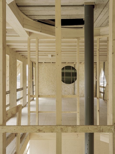  Ein Blick in den Rohbau des Holzgebäudes, aufgenommen durch die Pfosten-Riegel-Konstruktion der Holzrahmen. Man schaut durch das Gitterwerk der vertikalen und horizontalen Holzbalken auf den dahinter liegenden Raum. Die Deckenbalken sind freigelegt. Ein großes, schwarzes Kunststoffrohr verläuft vertikal auf der rechten Seite. Im Hintergrund ist eine runde Fensteröffnung in der mit OSB-Platten verkleideten Wand zu sehen.