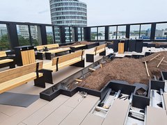 „Baustelle einer modernen Dachterrasse mit stufenförmig angeordneten Holzbänken, frisch angelegten Pflanzbeeten und Blick auf städtische Hochhäuser hinter einem Glasgeländer.“