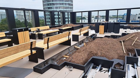 „Baustelle einer modernen Dachterrasse mit stufenförmig angeordneten Holzbänken, frisch angelegten Pflanzbeeten und Blick auf städtische Hochhäuser hinter einem Glasgeländer.“