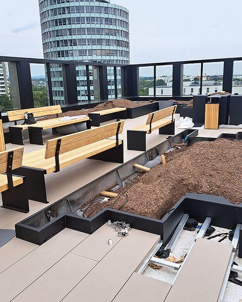 „Baustelle einer modernen Dachterrasse mit stufenförmig angeordneten Holzbänken, frisch angelegten Pflanzbeeten und Blick auf städtische Hochhäuser hinter einem Glasgeländer.“