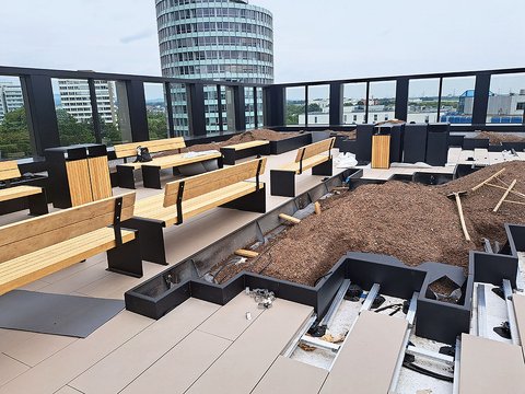 „Baustelle einer modernen Dachterrasse mit stufenförmig angeordneten Holzbänken, frisch angelegten Pflanzbeeten und Blick auf städtische Hochhäuser hinter einem Glasgeländer.“