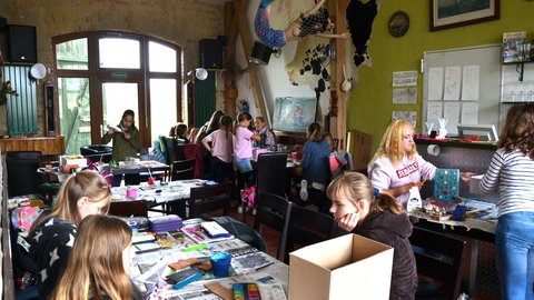 Eine Weitwinkelaufnahme eines belebten, gemütlichen Raumes mit vielen Tischen, an denen überwiegend Mädchen an verschiedenen Kunstprojekten arbeiten. Die Tische sind mit Zeitungspapier, Farbkästen, Pinseln und Bastelmaterial bedeckt. Im Vordergrund sitzt ein Mädchen mit langen braunen Haaren und malt. An der hinteren Wand hängt ein ausgefallenes Kunstwerk: eine als Meerjungfrau gestaltete Schaufensterpuppe in einem Fischnetz.