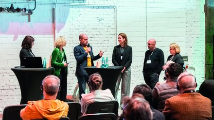 Podiumsdiskussion vor Publikum in Industriehalle Mehrere Sprecher:innen diskutieren auf einer Bühne vor sitzendem Publikum.