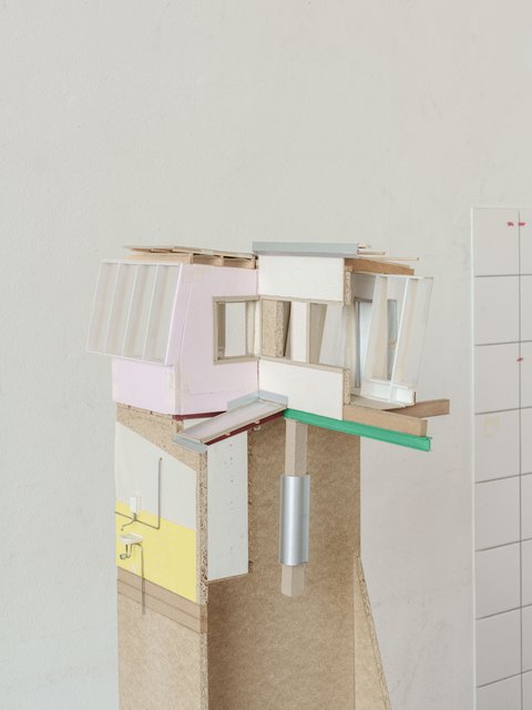  Eine Seitenansicht des zweiten Modells, die die Schichtung der Materialien und die komplexen räumlichen Beziehungen weiter verdeutlicht. Man sieht die gläserne Fassade, die verschiedenen Wandfarben (rosa und gelb), die Holzkonstruktion und die Metalldetails. Der Sockel ist aus Spanplatten und ragt hoch auf. Der Hintergrund ist wiederum die weiße Wand mit dem gefliesten Bereich.