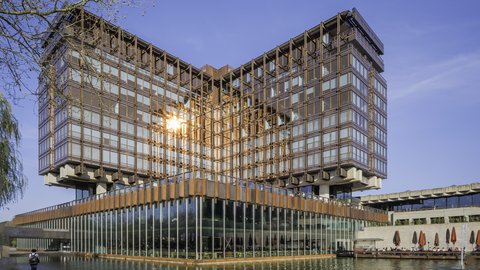 Großer moderner Bau mit rostfarbenem Fassadenraster über einem verglasten Sockel, umgeben von Wasser; Weg und Café‑Terrasse mit Schirmen.