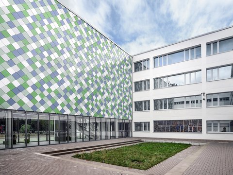Innenhof einer modernen Schule mit mehrstöckigem Gebäude und Fassade aus quadratischen, farblich gemusterten Paneelen neben einem weißen Gebäudeteil mit vielen Fenstern