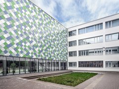 Innenhof einer modernen Schule mit mehrstöckigem Gebäude und Fassade aus quadratischen, farblich gemusterten Paneelen neben einem weißen Gebäudeteil mit vielen Fenstern