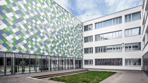 Innenhof einer modernen Schule mit mehrstöckigem Gebäude und Fassade aus quadratischen, farblich gemusterten Paneelen neben einem weißen Gebäudeteil mit vielen Fenstern