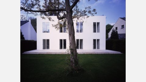"Zweigeschossiges modernes Wohnhaus mit schneeweißer Fassade. Symmetrisch angeordnete große Fenster in vier Reihen. Breite gepflasterte Terrasse im Vordergrund. Ein großer knorriger Baum mit ausladendem Ast vor der Hausfassade. Grüner Rasen, umgeben von Hecken. Blauer Himmel."