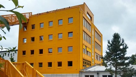 Außenansicht eines gelb-orangenen Gebäudes; davorstehende Personengruppe betrachtet das Gebäude