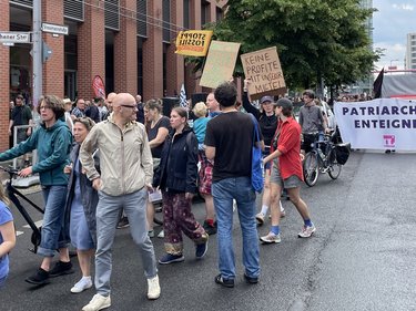Das Bild zeigt eine politische Demonstration auf einer breiten Stadtstraße bei Tageslicht. Viele Menschen gehen dicht nebeneinander, einige schieben Fahrräder. Die Straße ist nass, als hätte es kurz zuvor geregnet. Die Teilnehmenden tragen Alltagskleidung wie Jacken, Jeans und Rucksäcke. Mehrere Personen halten handgemalte Protestschilder hoch. Darauf stehen unter anderem die Slogans „Keine Profite mit unserer Miete“, „Miet-Haie zu Fischstäbchen“ und „Stoppt fossile Subventionen“. Weiter hinten ist ein großes Banner mit der Aufschrift „Patriarchat enteignen“ zu sehen. Im Hintergrund stehen Backsteingebäude, Bäume und Straßenschilder an einer Kreuzung. Die Atmosphäre wirkt engagiert, friedlich und politisch.