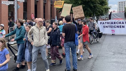 Das Bild zeigt eine politische Demonstration auf einer breiten Stadtstraße bei Tageslicht. Viele Menschen gehen dicht nebeneinander, einige schieben Fahrräder. Die Straße ist nass, als hätte es kurz zuvor geregnet. Die Teilnehmenden tragen Alltagskleidung wie Jacken, Jeans und Rucksäcke. Mehrere Personen halten handgemalte Protestschilder hoch. Darauf stehen unter anderem die Slogans „Keine Profite mit unserer Miete“, „Miet-Haie zu Fischstäbchen“ und „Stoppt fossile Subventionen“. Weiter hinten ist ein großes Banner mit der Aufschrift „Patriarchat enteignen“ zu sehen. Im Hintergrund stehen Backsteingebäude, Bäume und Straßenschilder an einer Kreuzung. Die Atmosphäre wirkt engagiert, friedlich und politisch.