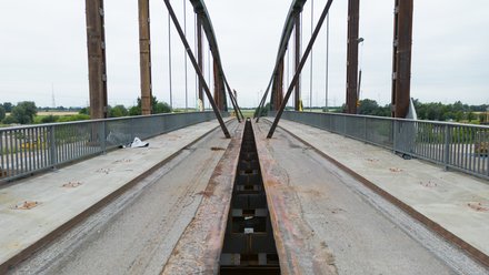 Blick über die unfertige Brücke: zwei hohe Stahlbögen und Geländer, mittig ein offener rostiger Träger; Betonränder ohne Belag, schräge Montage-Streben verbinden Bogen und Deck.