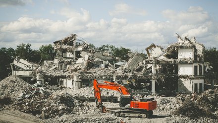 Abrissbaustelle: orange Bagger vor großen Schutthalden und Resten eines eingestürzten Betongebäudes; Bäume und Wolkenhimmel im Hintergrund.