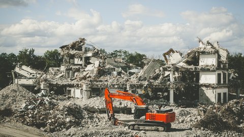 Abrissbaustelle: orange Bagger vor großen Schutthalden und Resten eines eingestürzten Betongebäudes; Bäume und Wolkenhimmel im Hintergrund.