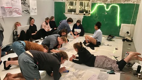  "Etwa 12 Personen knien und liegen auf dem Boden eines hell erleuchteten Ausstellungsraumes. Sie arbeiten an großen weißen Plänen mit botanischen Zeichnungen und Skizzen. An der Wand leuchtet eine grüne Neonskulptur. Schuhe und Taschen liegen verstreut umher."