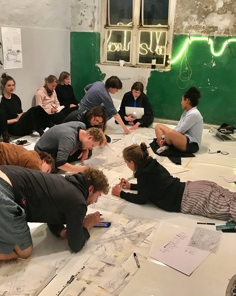  "Etwa 12 Personen knien und liegen auf dem Boden eines hell erleuchteten Ausstellungsraumes. Sie arbeiten an großen weißen Plänen mit botanischen Zeichnungen und Skizzen. An der Wand leuchtet eine grüne Neonskulptur. Schuhe und Taschen liegen verstreut umher."