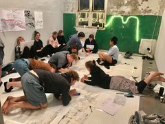  "Etwa 12 Personen knien und liegen auf dem Boden eines hell erleuchteten Ausstellungsraumes. Sie arbeiten an großen weißen Plänen mit botanischen Zeichnungen und Skizzen. An der Wand leuchtet eine grüne Neonskulptur. Schuhe und Taschen liegen verstreut umher."